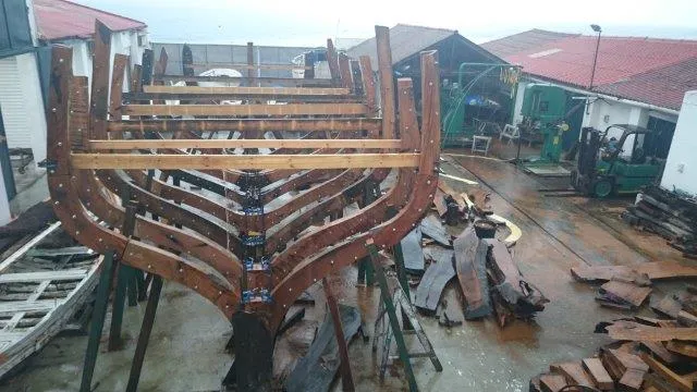 Estructura del casco de un barco de madera en construcción en un astillero tradicional, con cuadernas curvas apoyadas sobre soportes metálicos y tablones de madera esparcidos por el suelo húmedo, con edificios de tejados rojos al fondo.