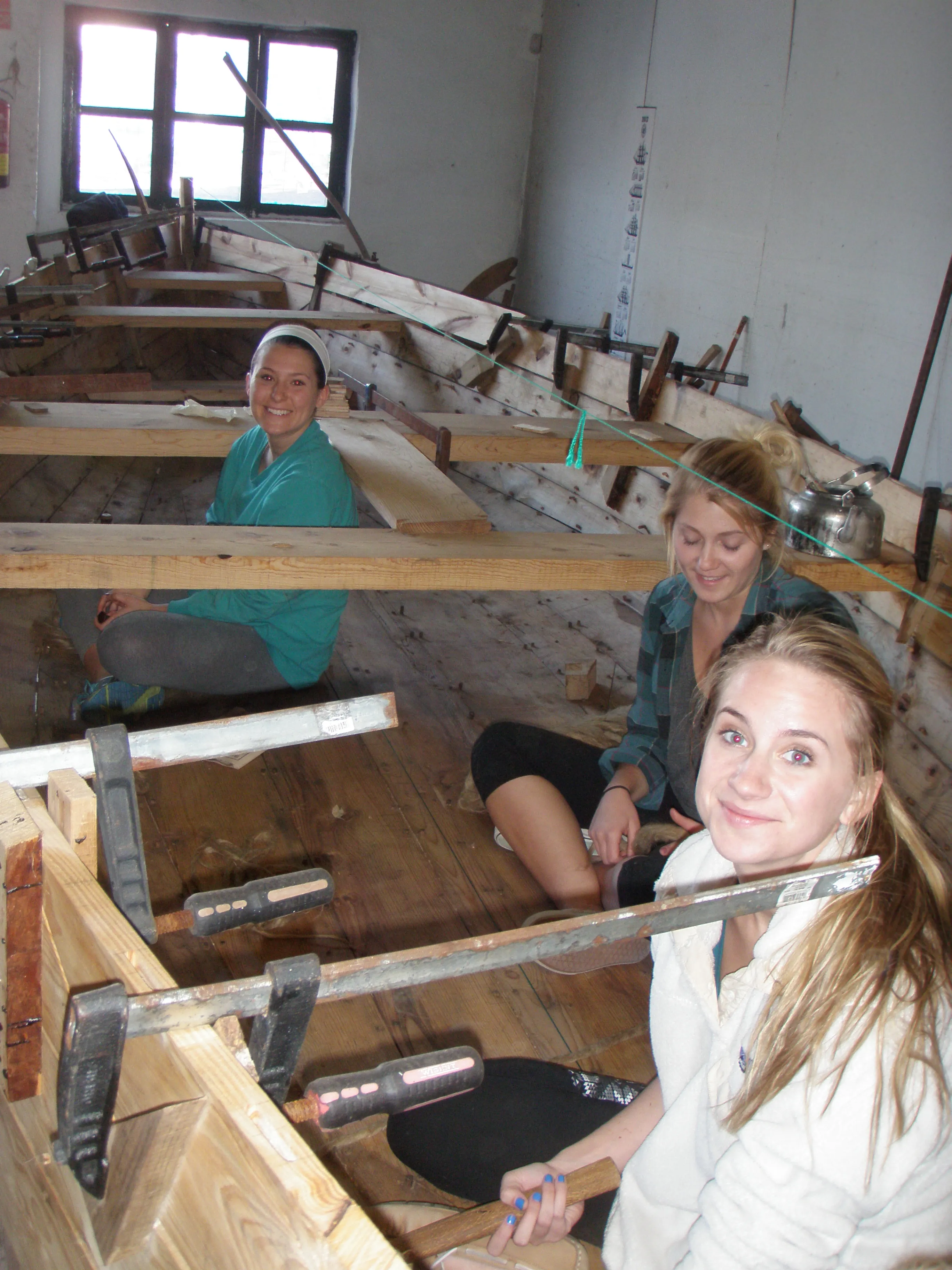 Tres mujeres sentadas dentro del casco de un barco de madera en construcción, rodeadas de abrazaderas metálicas y herramientas de carpintería en un taller con una ventana al fondo.