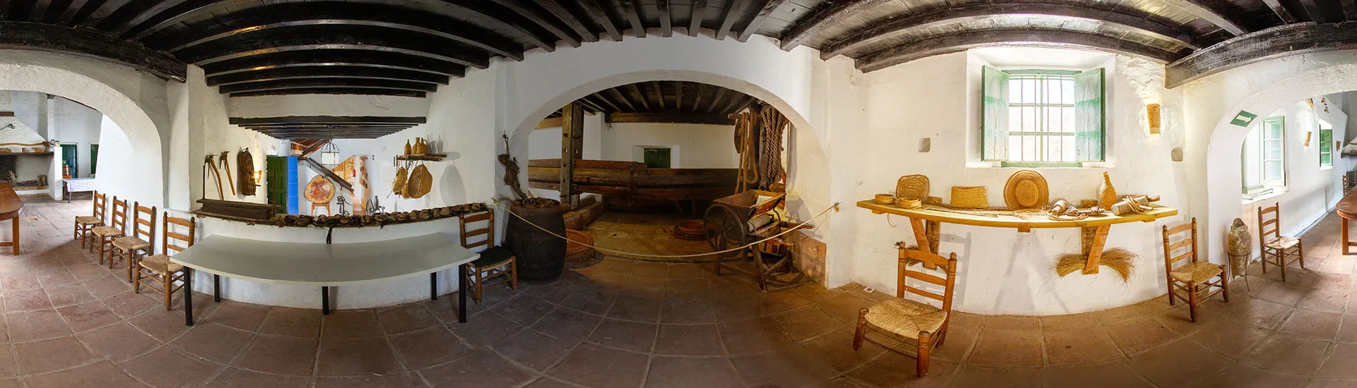 Vista panorámica del interior de una habitación con paredes blancas, vigas de madera oscura en el techo, puertas arqueadas y sillas de madera dispuestas alrededor de las mesas.