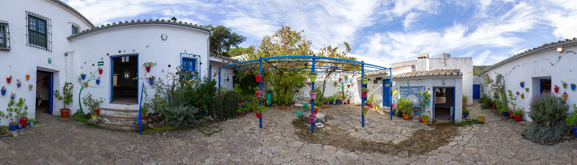 Vista panorámica de un patio soleado con edificios encalados, una pérgola de madera azul y coloridas decoraciones de cerámica en las paredes.
