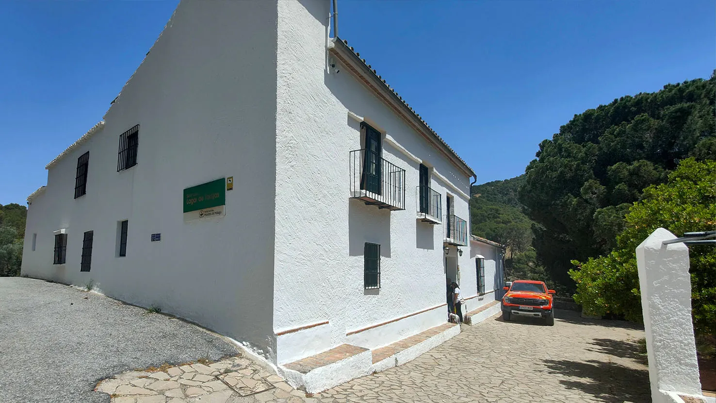 Edificio rural blanco de dos plantas con contraventanas verdes y una placa verde en la pared, un coche naranja aparcado cerca y una colina boscosa al fondo bajo un cielo azul.