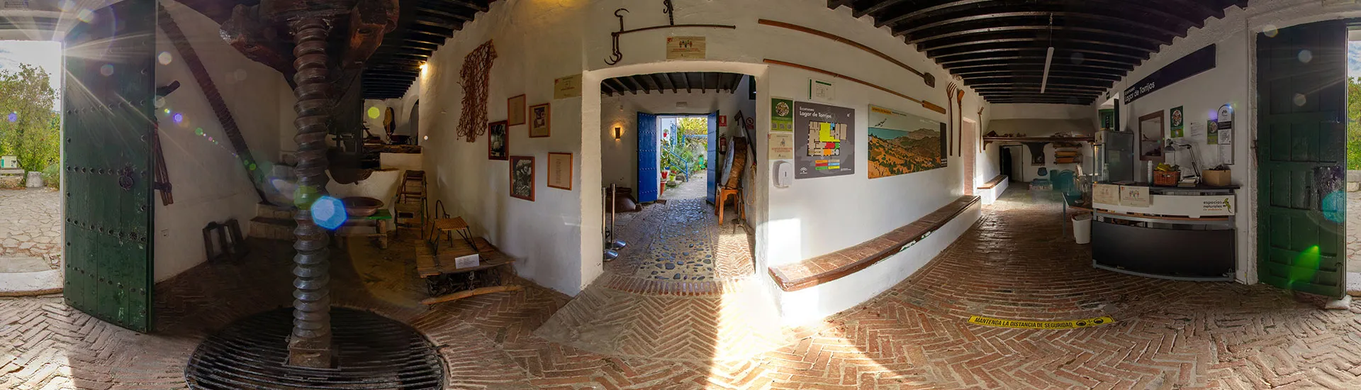 Interior panorámico de una tradicional sala de prensa vinícola con una gran prensa de tornillo de madera, paredes blancas arqueadas, cuadros y un suelo de adoquines que conduce a una puerta exterior.