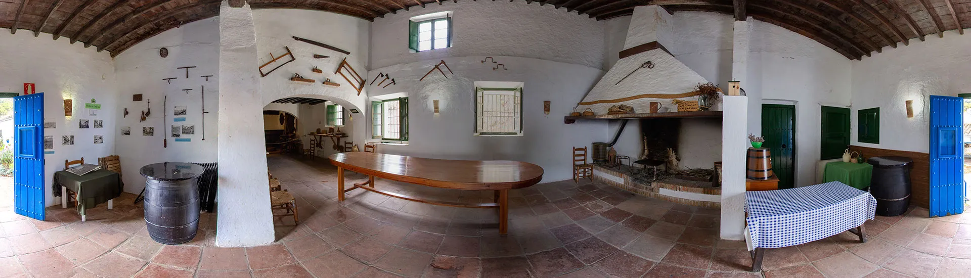 Vista panorámica interior de una habitación tradicional con paredes blancas, suelo de ladrillo, vigas de madera en el techo, gran chimenea, mesa larga de madera, barriles y puertas azules