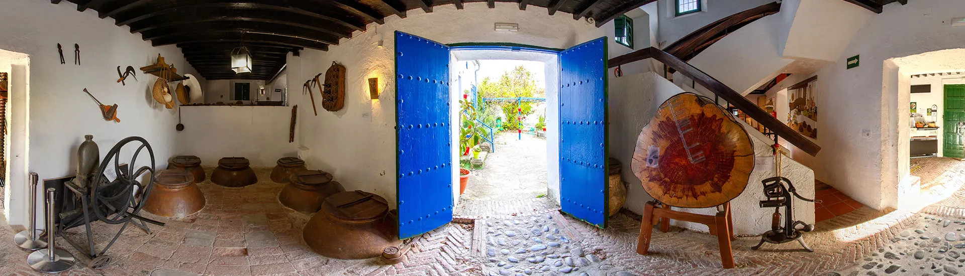 Interior panorámico de una prensa de vino tradicional con grandes puertas dobles azules que dan a un patio, paredes blancas con vigas de madera oscura en el techo, vasijas de barro sobre un suelo de piedra y maquinaria de prensado antigua que incluye un gran mecanismo de rueda de madera