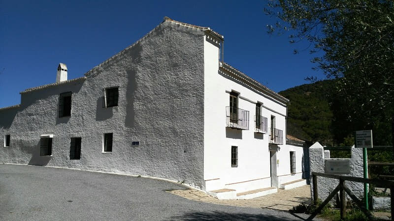Edificio tradicional de dos plantas encalado, con marcos de ventana oscuros, chimenea y un pequeño balcón con barandilla de hierro, sobre un cielo azul despejado y con un patio de grava al frente.