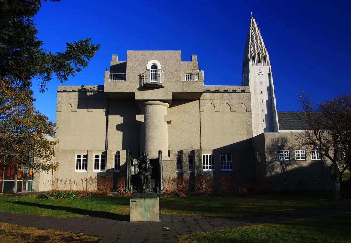 Zewnętrze Muzeum Einara Jónssona prezentujące betonowy budynek z cylindryczną wieżą i rzeźbą z brązu w ogrodzie na pierwszym planie, z widoczną w tle wysoką iglicą kościoła Hallgrímskirkja na tle głębokiego, błękitnego nieba.