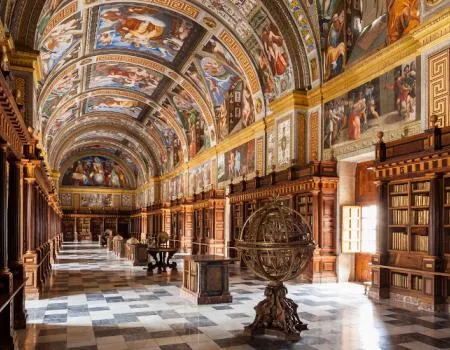 Ozdobne wnętrze biblioteki ze sklepieniem ozdobionym freskami, drewnianymi regałami wzdłuż ścian i dużą kulą niebieską pośrodku.