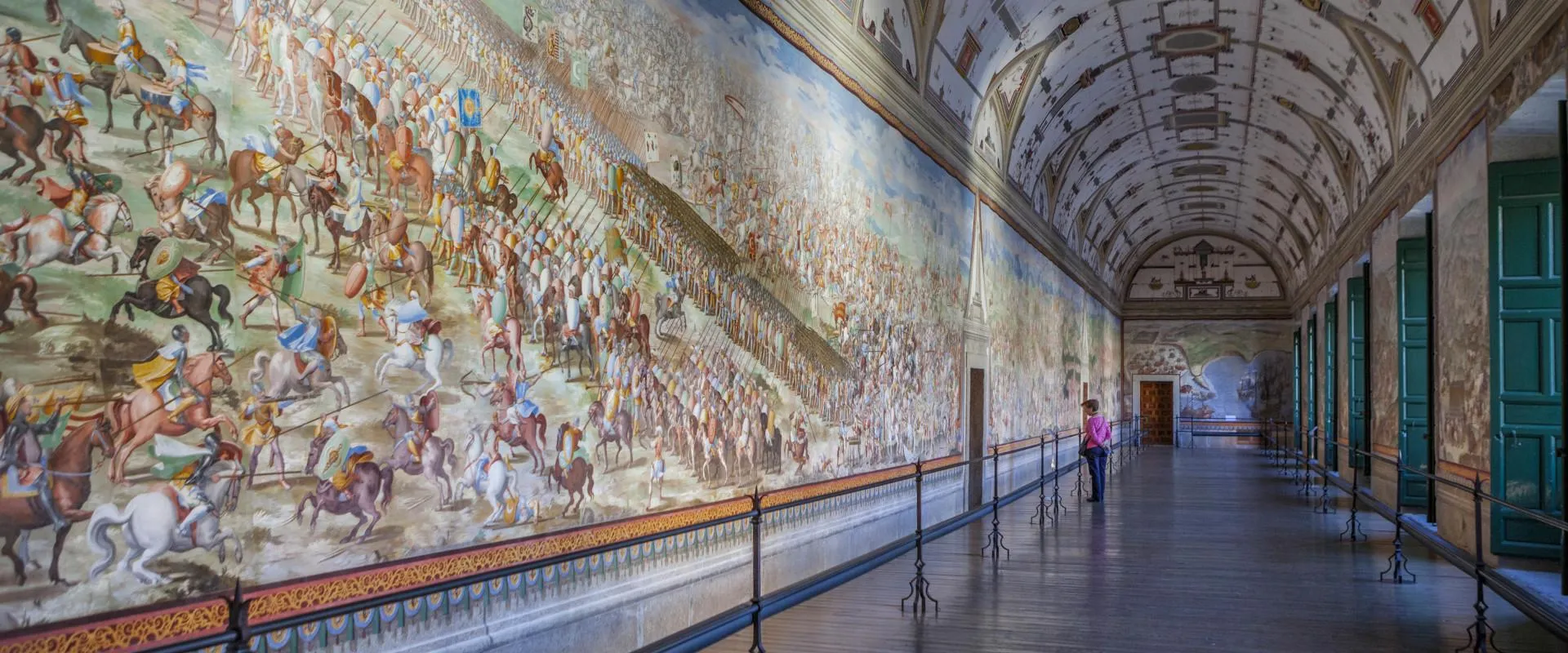 Long gallery with a large mural depicting a battle scene, green doors, and a visitor walking through