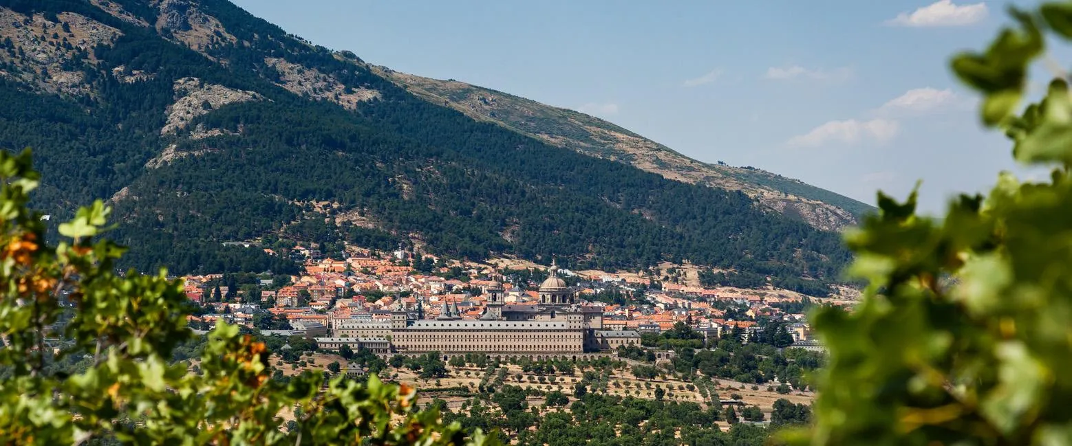 Panoramic view of the Royal Site of San Lorenzo de El Escorial with buildings surrounded by green hills and trees in the foreground