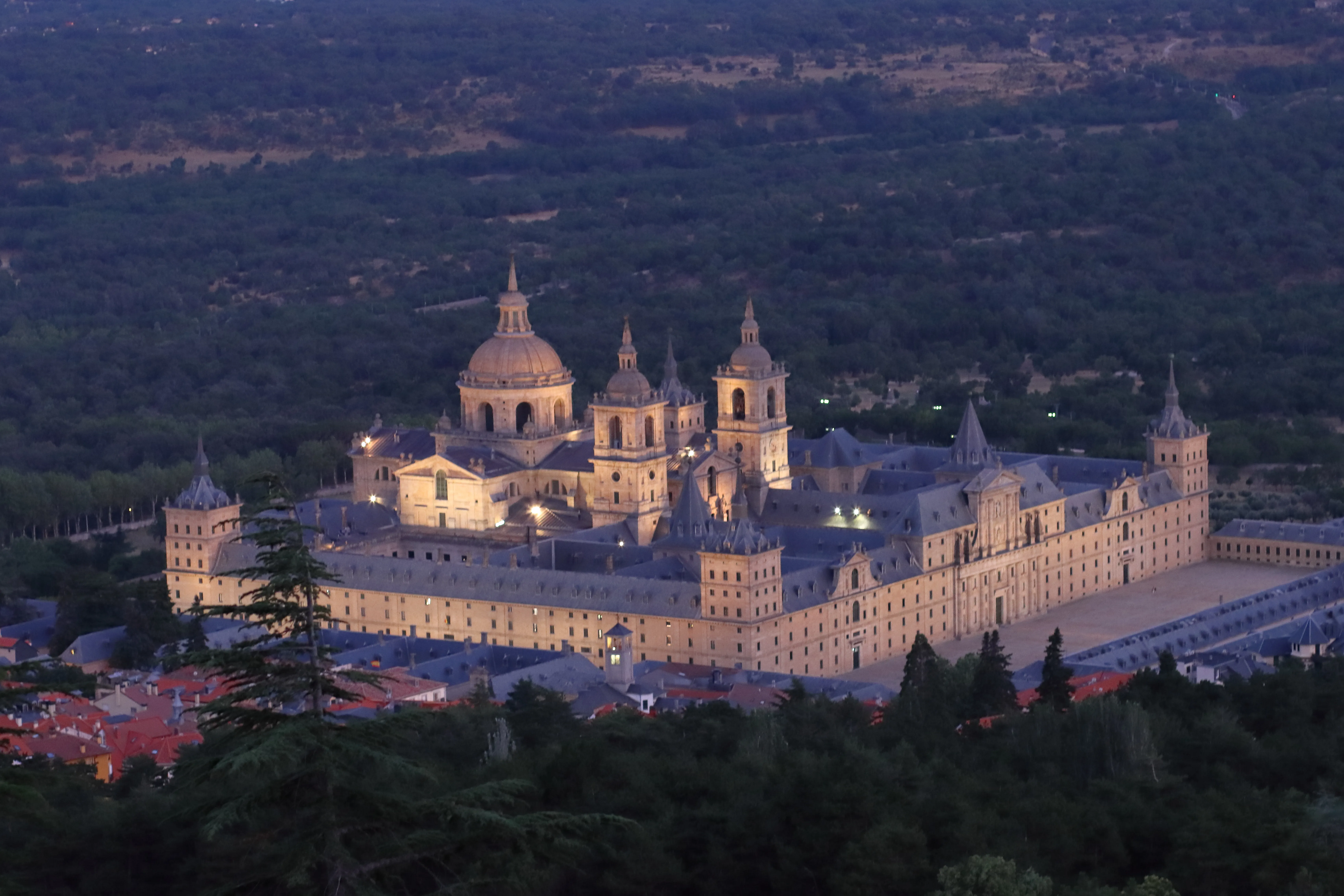 Aerial night view of the illuminated Royal Site of San Lorenzo de El Escorial with its domes and towers lit against a dark forested background
