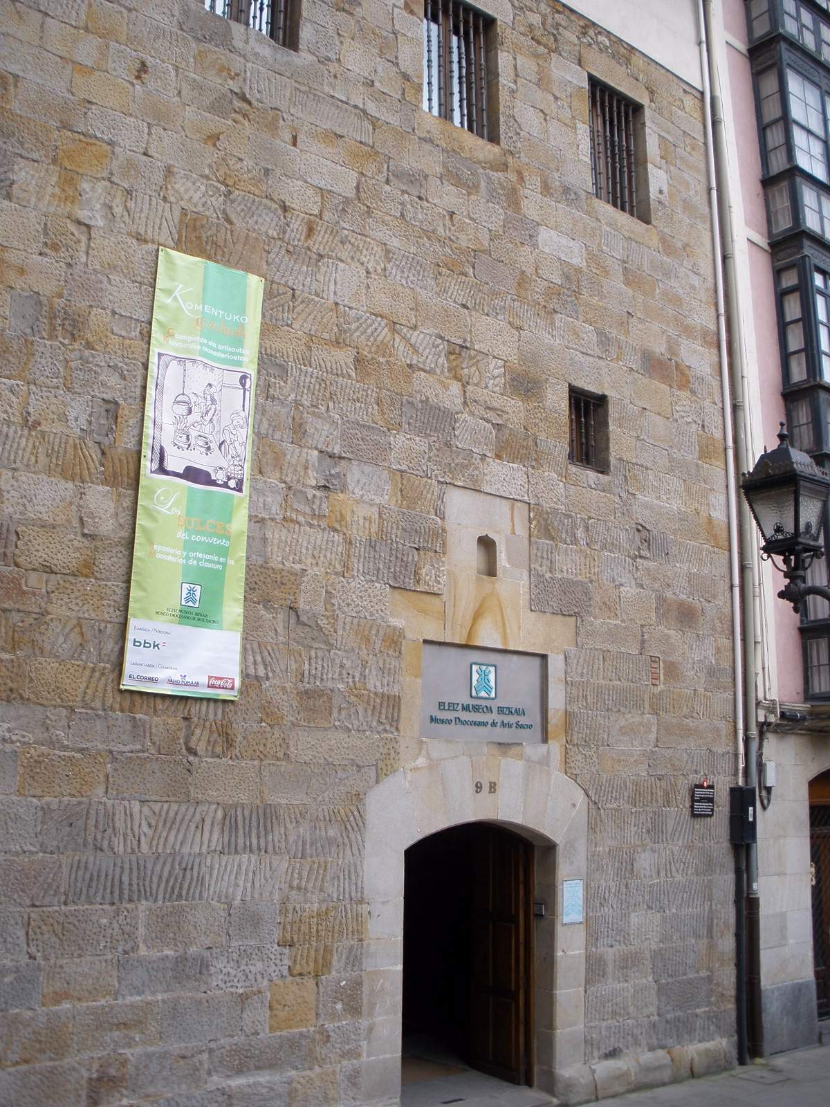 Kamienna fasada Eleiz Museoa z łukowym wejściem, szyldem muzeum oraz zielonym banerem wystawowym zawieszonym na ścianie