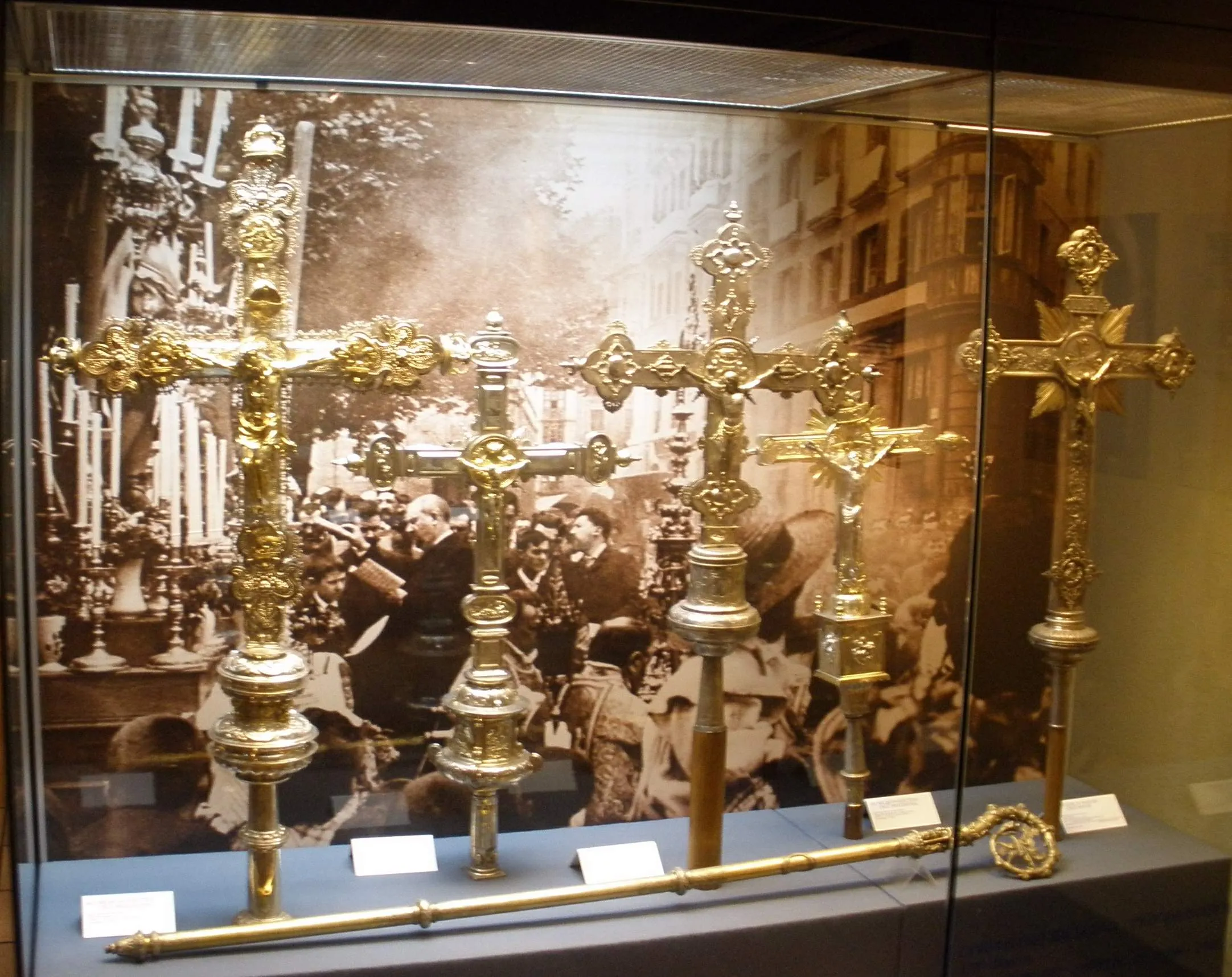 Pięć złotych krzyży procesyjnych i długa laska wyeksponowane w szklanej gablocie muzealnej na tle zdjęcia w sepii przedstawiającego procesję religijną