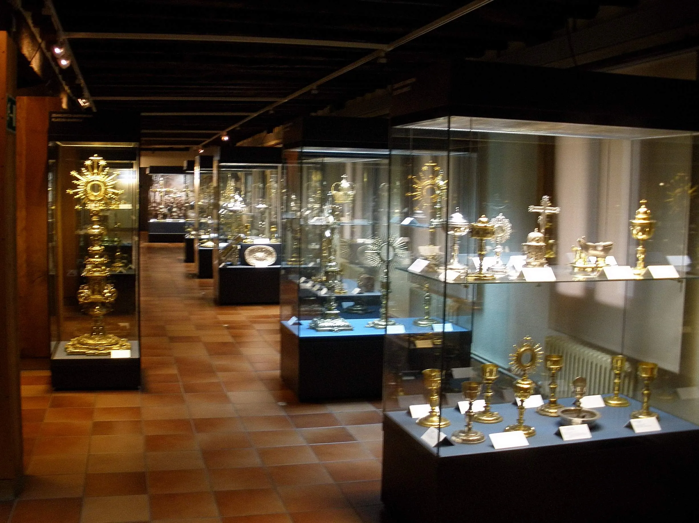 Wnętrze muzeum z rzędami szklanych gablot zawierających złote artefakty religijne, takie jak kielichy i monstrancje, ustawionych na podłodze z terakoty pod ciemnym belkowanym sufitem.