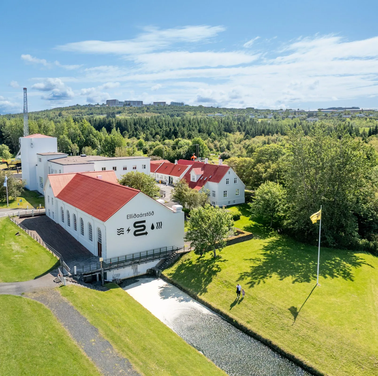 Luchtfoto van witte gebouwen met rode daken bij de waterkrachtcentrale Elliðaárstöð, omringd door groene bomen en een waterkanaal.