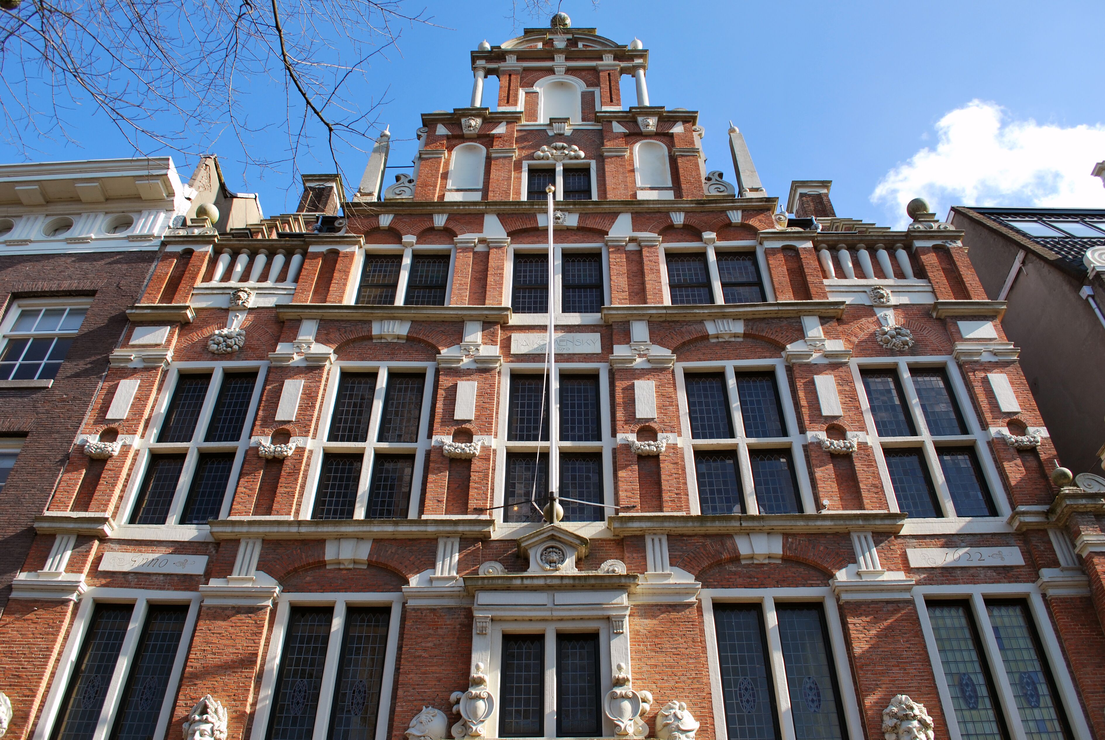 Fachada de ladrillo rojo de un edificio histórico con adornos blancos ornamentados, ventanas altas y estrechas, cantería decorativa y un frontón escalonado bajo un cielo azul claro.