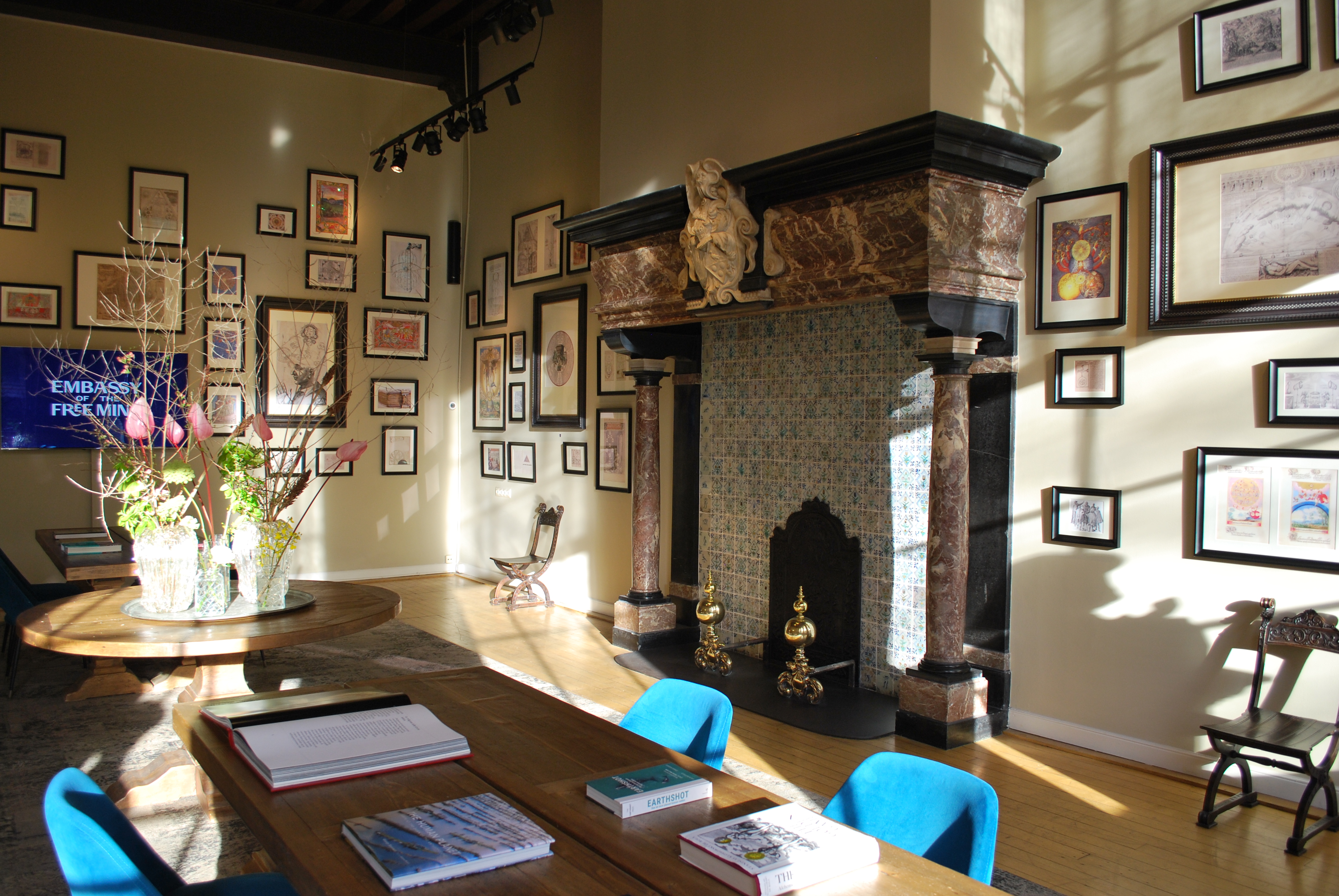 Habitación interior con luz solar en la Embassy of the Free Mind con chimenea ornamentada, obras de arte enmarcadas en las paredes, mesas con libros y sillas azules.