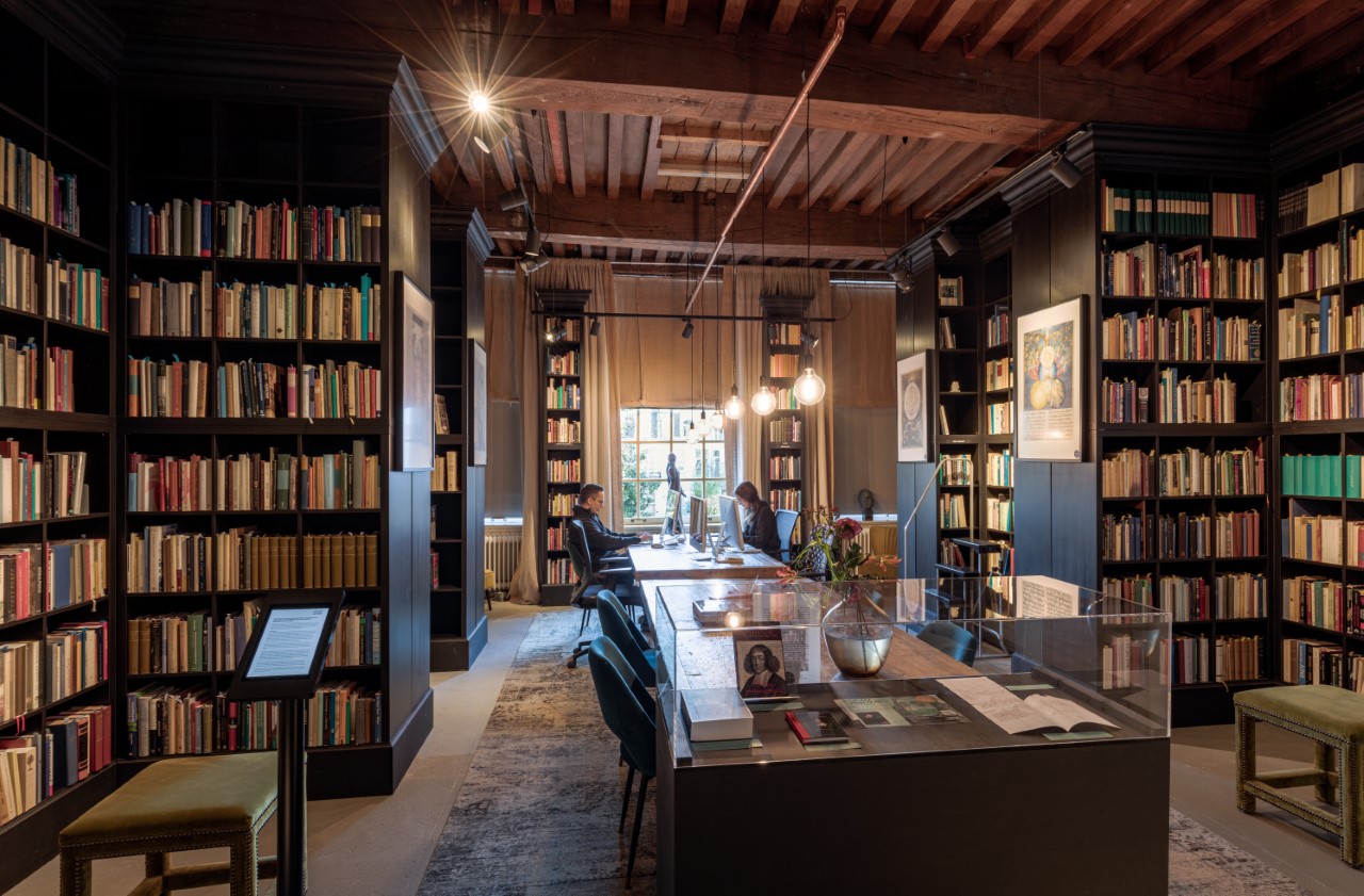 Interior de una biblioteca con altas estanterías, una mesa central con una vitrina de cristal y vigas de madera en el techo