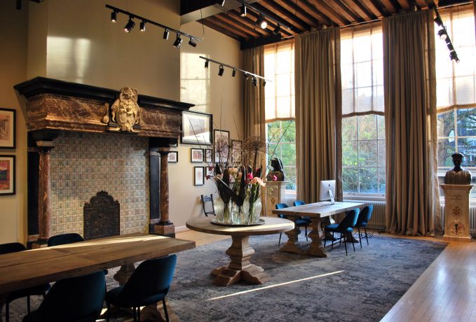 Elegante habitación interior con chimenea ornamentada, vigas de madera en el techo, ventanales altos con cortinas, mesas, sillas y obras de arte enmarcadas en las paredes