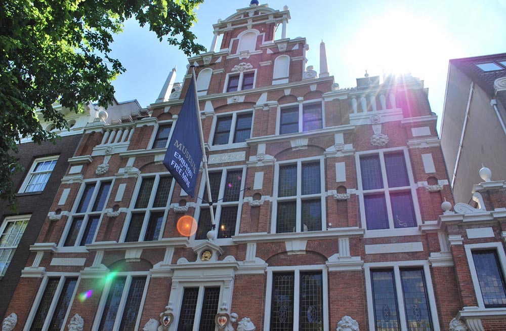 Edificio histórico de ladrillo rojo con molduras decorativas blancas y múltiples ventanas. Bandera azul con texto visible, sol creando destellos de lente, árboles en los bordes.