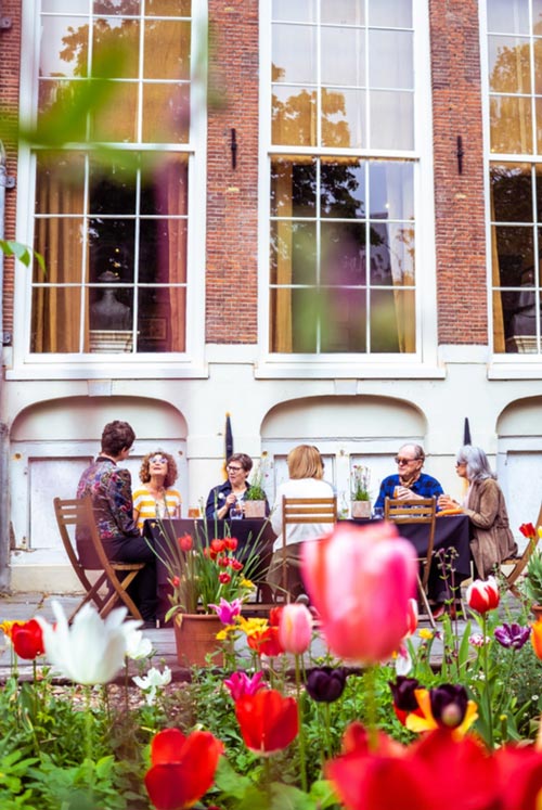 Fachada histórica de edificio de ladrillo con grandes ventanas y personas sentadas en mesas al aire libre en un vibrante jardín de tulipanes.