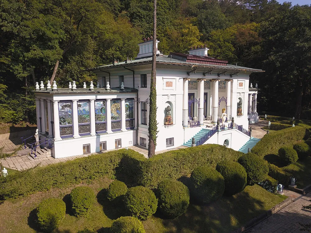 Villa blanca con columnas y ventanas decorativas rodeada de setos verdes y árboles.