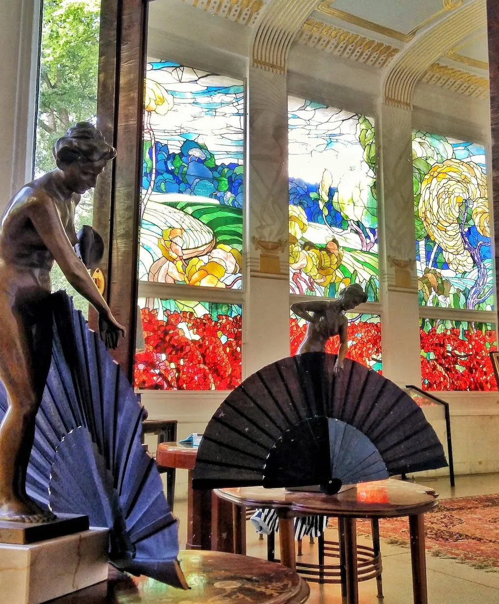 Escultura de bronce de una figura con un abanico, vidrieras con coloridas escenas surrealistas y una mesa con un abanico negro en una sala de arquitectura ornamentada