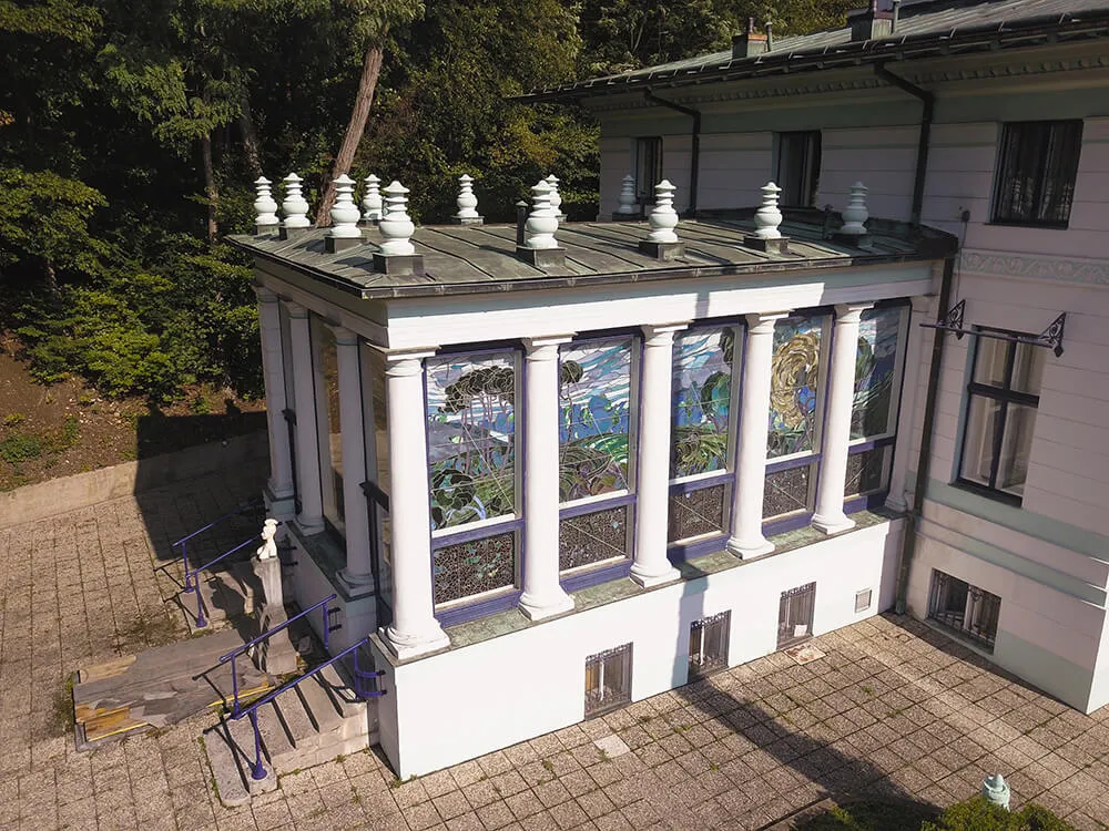 Vista aérea de una villa blanca con columnas y vidrieras, rodeada de árboles y un patio pavimentado