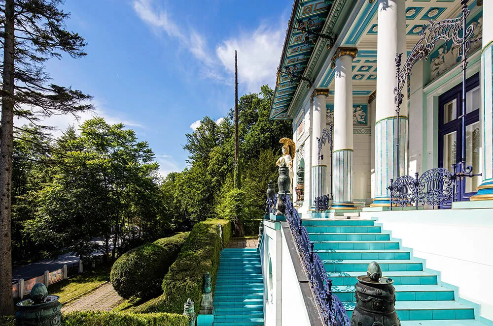Exterior del Museo Ernst Fuchs con escaleras turquesas, columnas blancas y barandillas decorativas contra un cielo azul