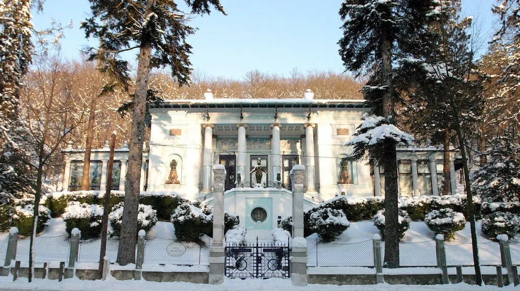 Villa cubierta de nieve con columnas clásicas y entrada central, rodeada de árboles y arbustos cargados de nieve