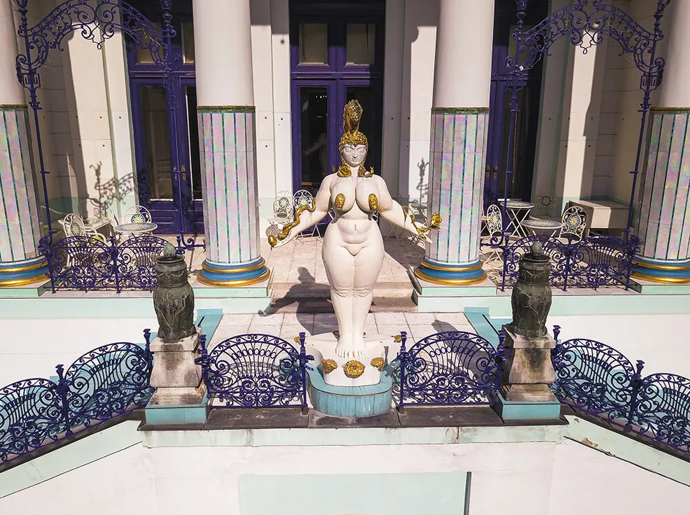 Una estatua blanca de una figura femenina con detalles dorados se encuentra en un patio con barandillas y columnas azules.