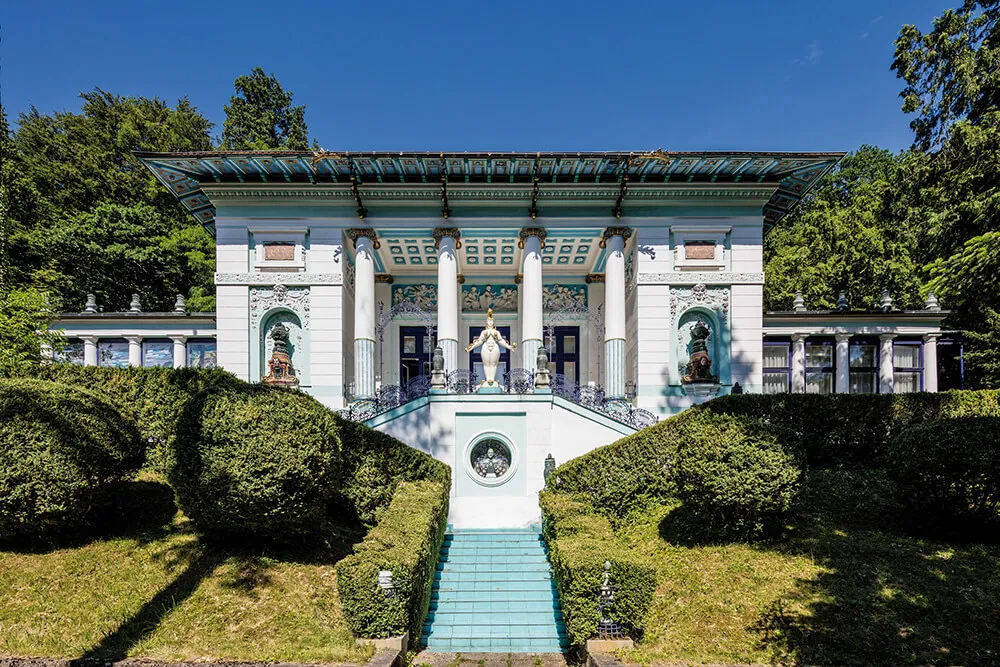 Muzeum Ernsta Fuchsa, biały budynek z ozdobnymi kolumnami i turkusowymi akcentami, otoczony zielonymi żywopłotami i turkusowymi schodami pod błękitnym niebem