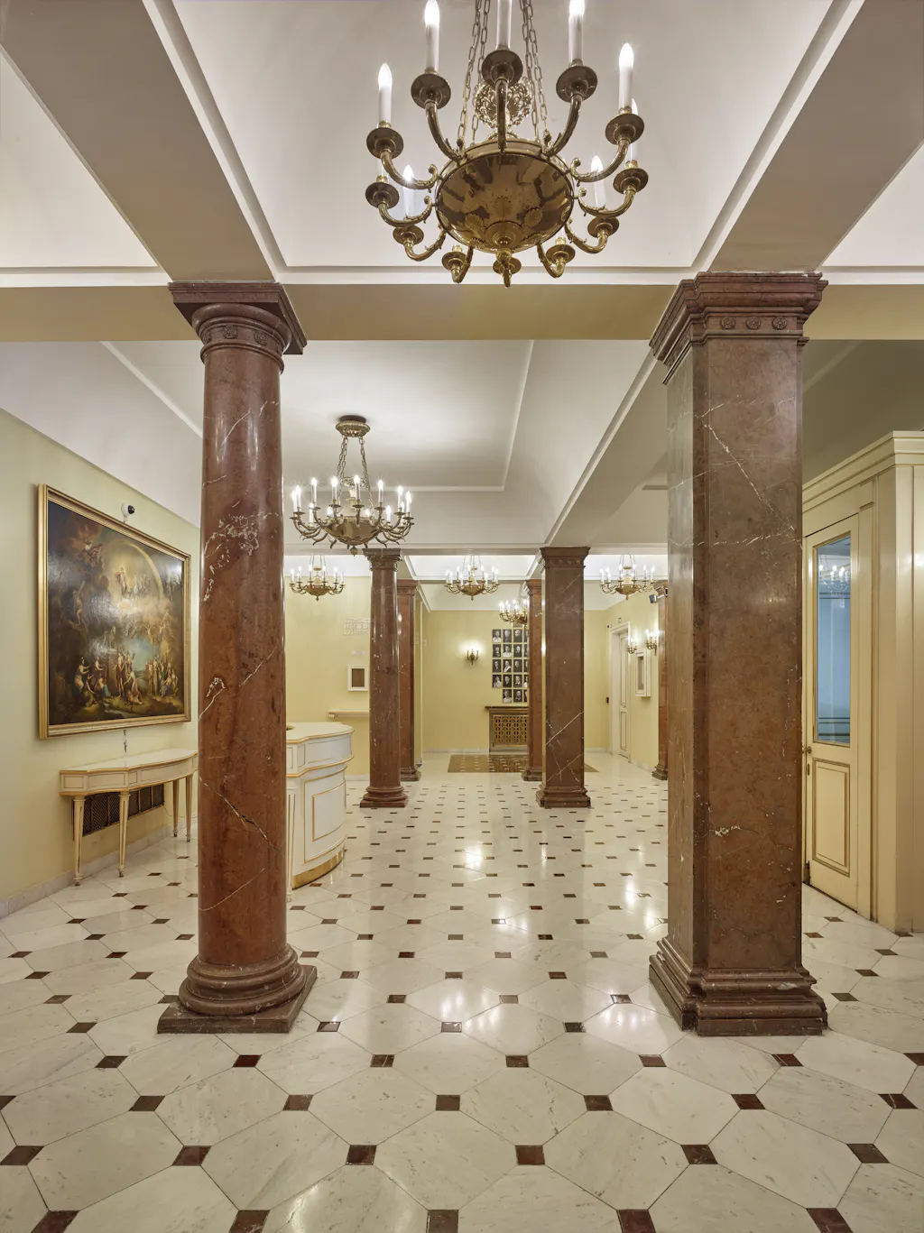 Ornate interior hallway with marble columns, chandeliers, and a patterned tile floor