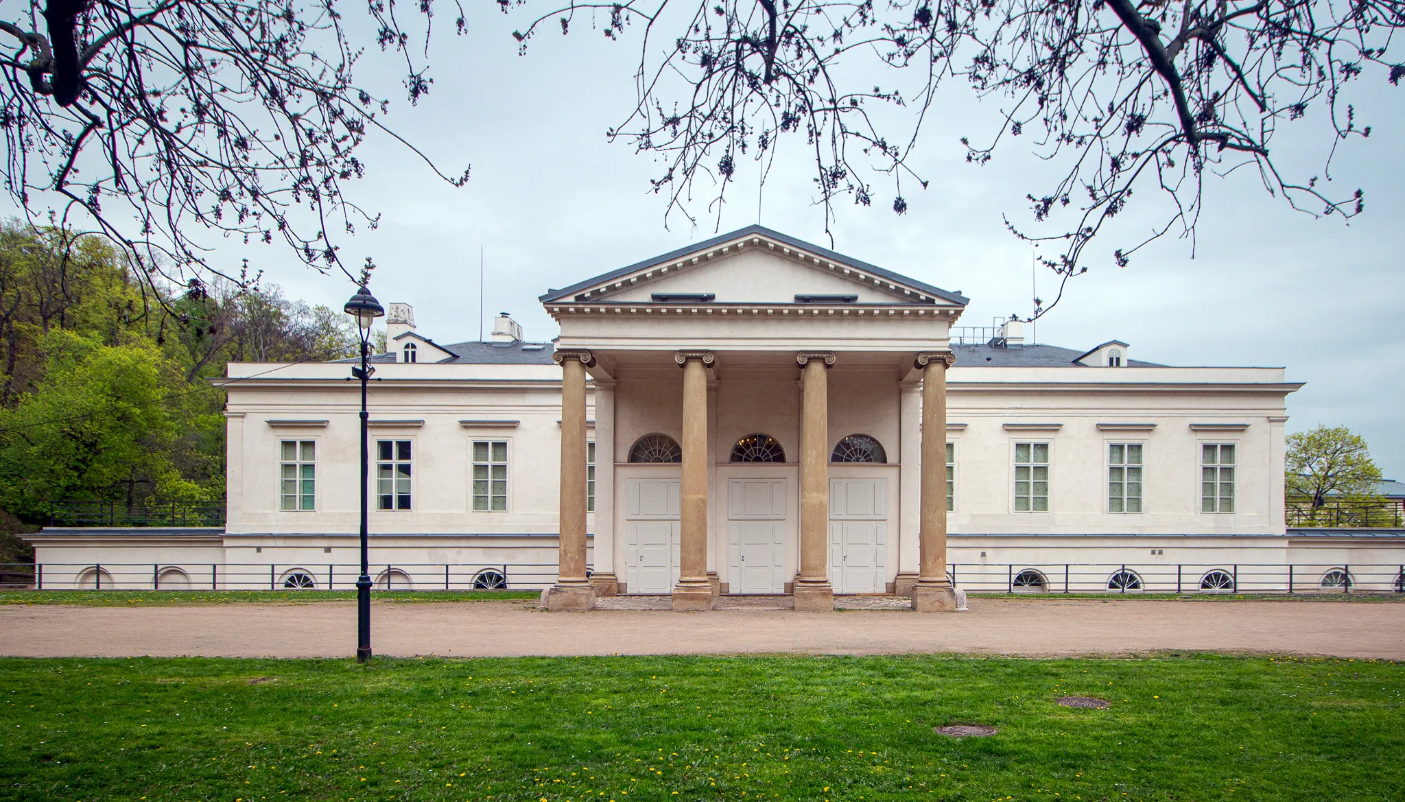 Edificio clásico blanco con columnas y un frontón triangular, rodeado de césped verde y árboles
