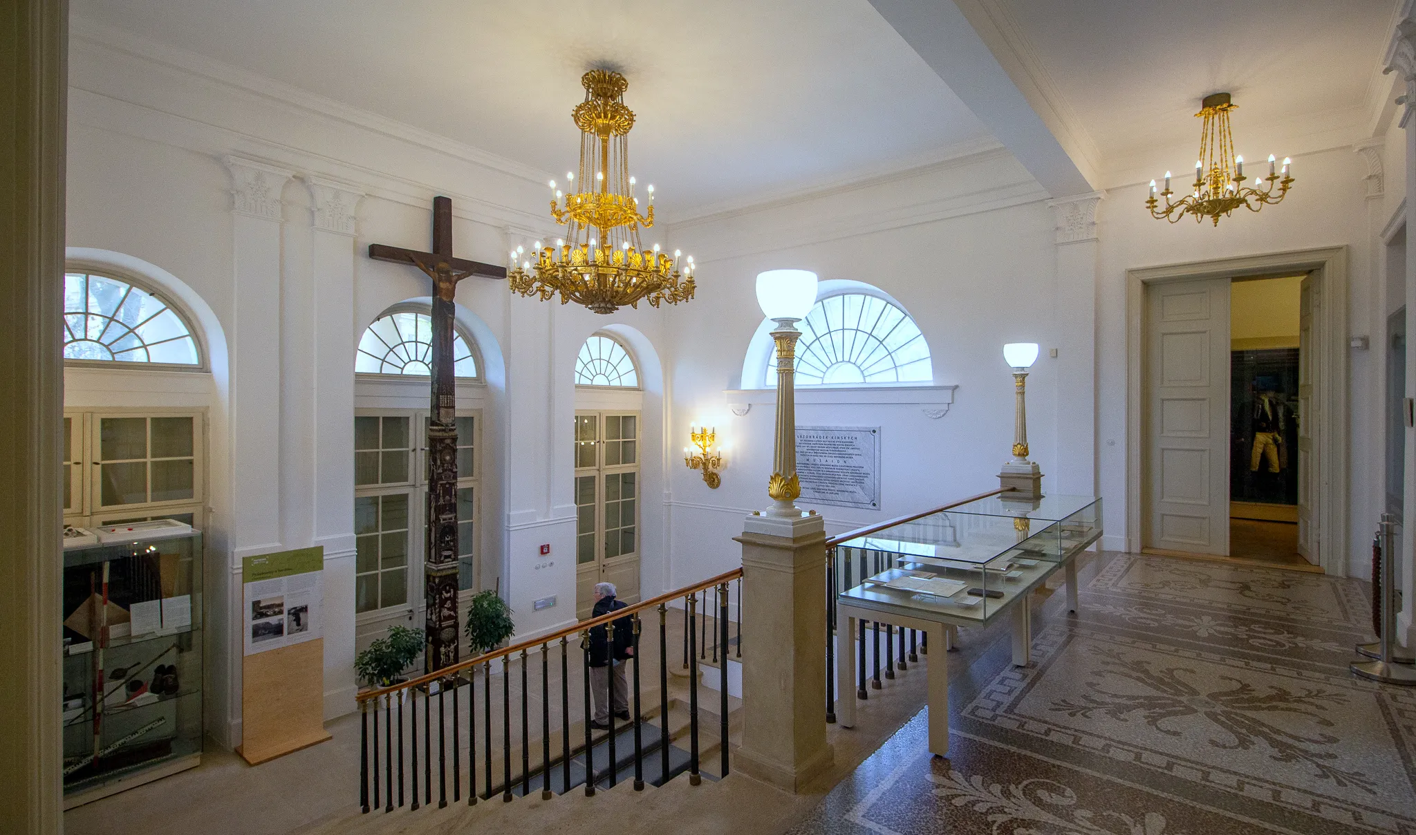 Salón interior con candelabros, ventanas arqueadas y una escalera con una vitrina de cristal