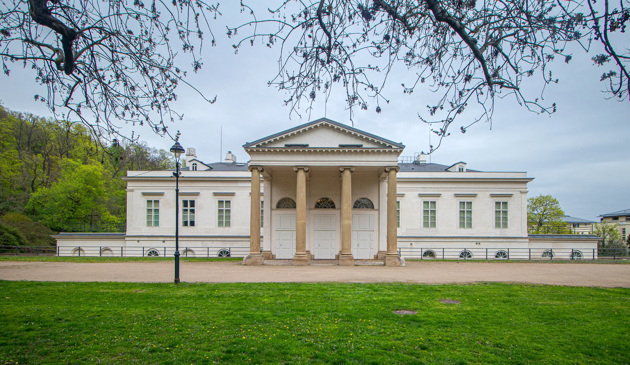 Edificio neoclásico blanco con columnas y un frontón triangular, rodeado de césped verde y árboles