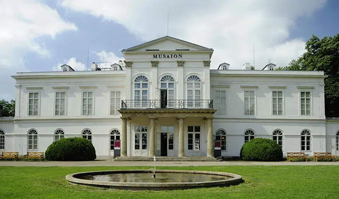 Edificio neoclásico blanco con la inscripción 'MUSÉUM' sobre la entrada, rodeado de césped verde y una fuente circular