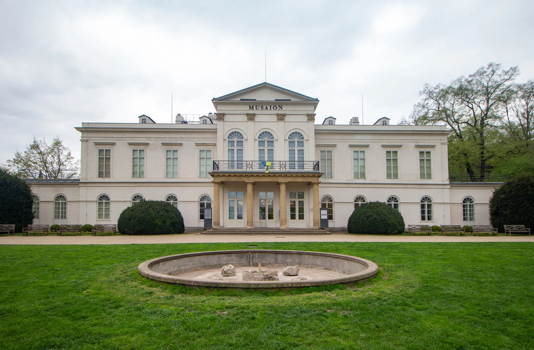 Edificio neoclásico blanco con columnas y ventanas arqueadas, rodeado de césped verde y árboles