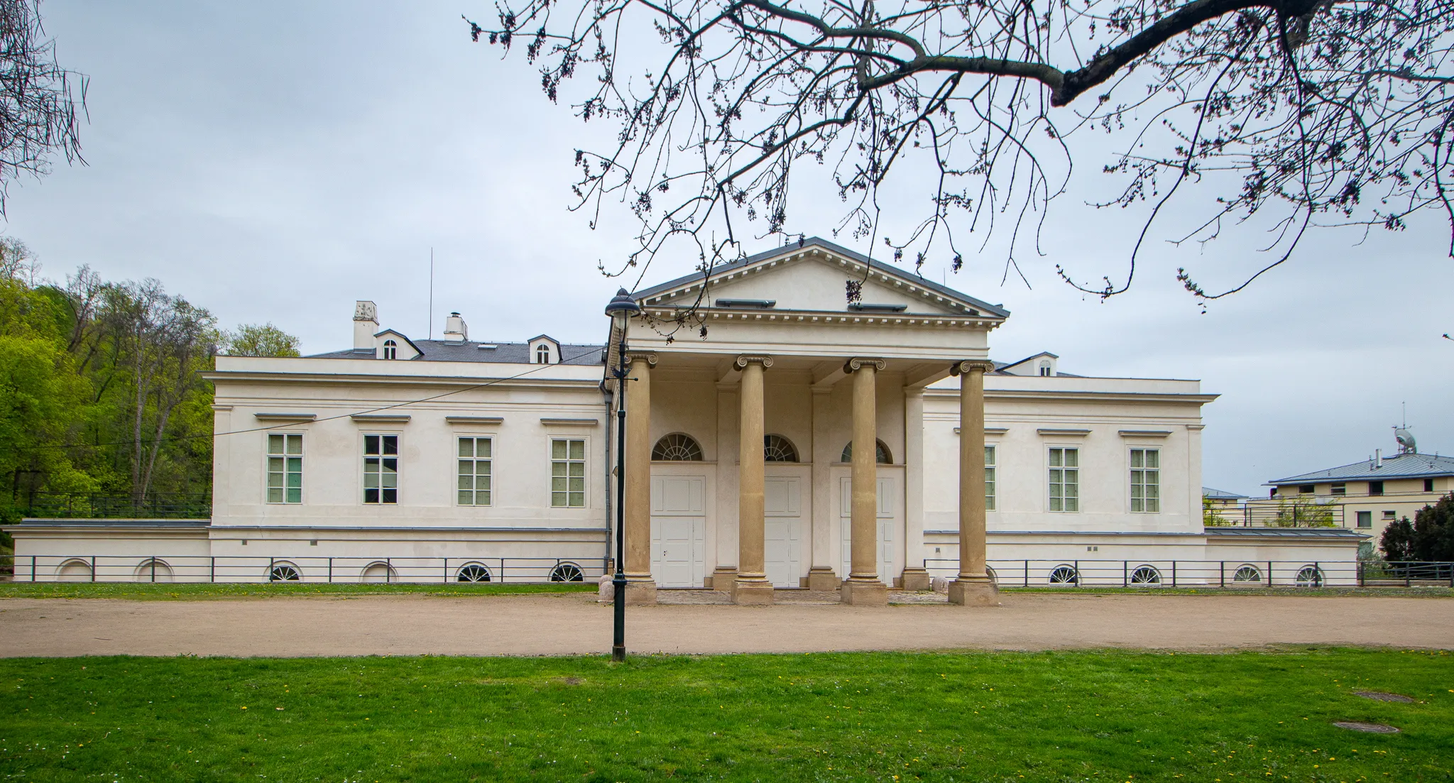 Edificio clásico blanco con un pórtico sostenido por columnas, césped verde en primer plano y ramas de árboles desnudas arriba