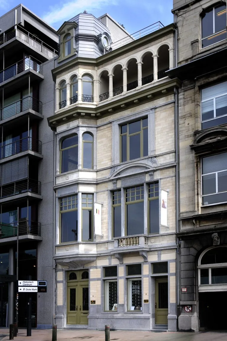 Edificio histórico de piedra de varias plantas con ventanas en arco, una torreta en esquina con cúpula y banderas en la fachada, flanqueado por edificios modernos
