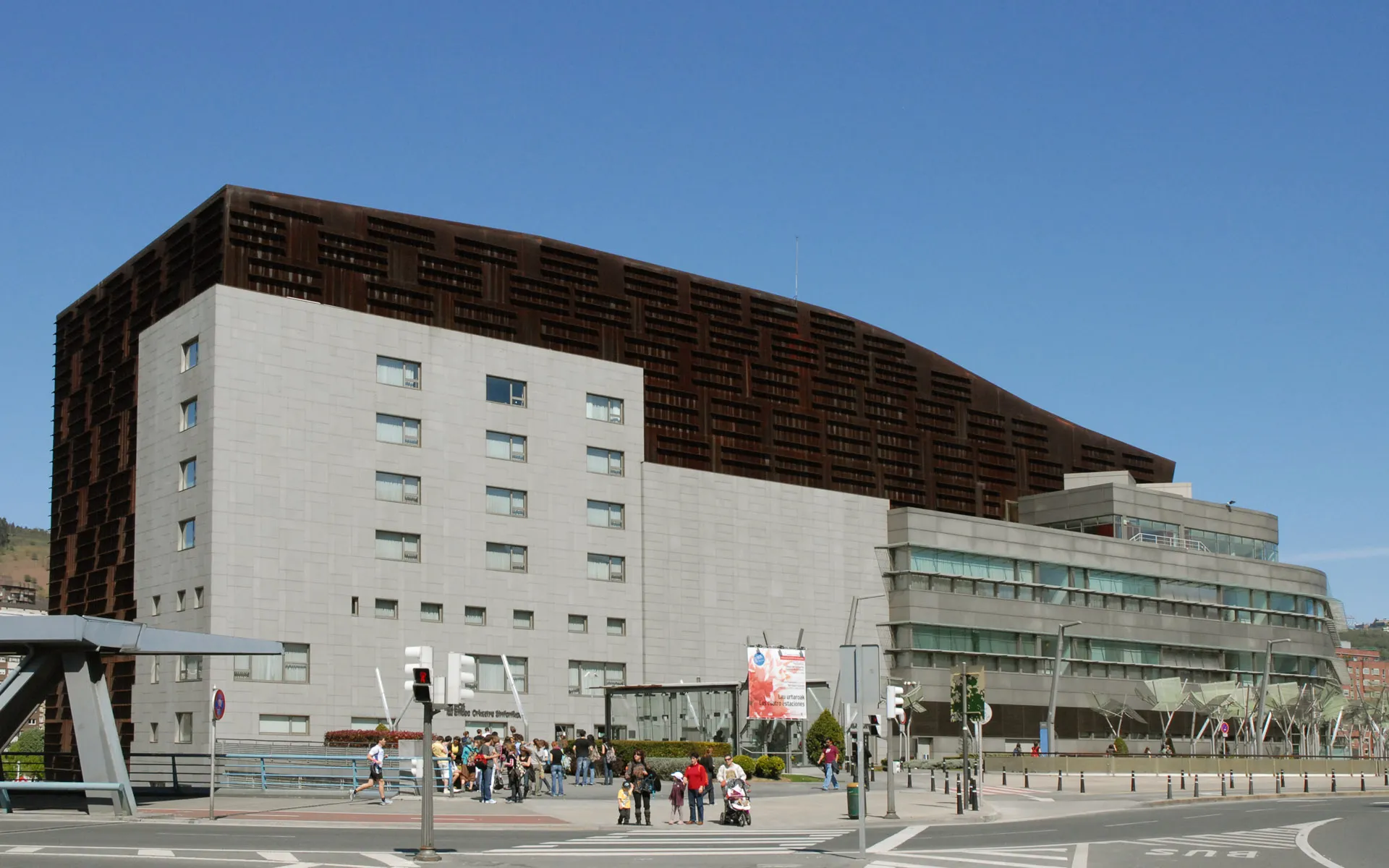 Widok z zewnątrz na Euskalduna Conference Centre w Bilbao, przedstawiający duży budynek z rdzawobrązową górną fasadą i białymi dolnymi partiami pod błękitnym niebem.