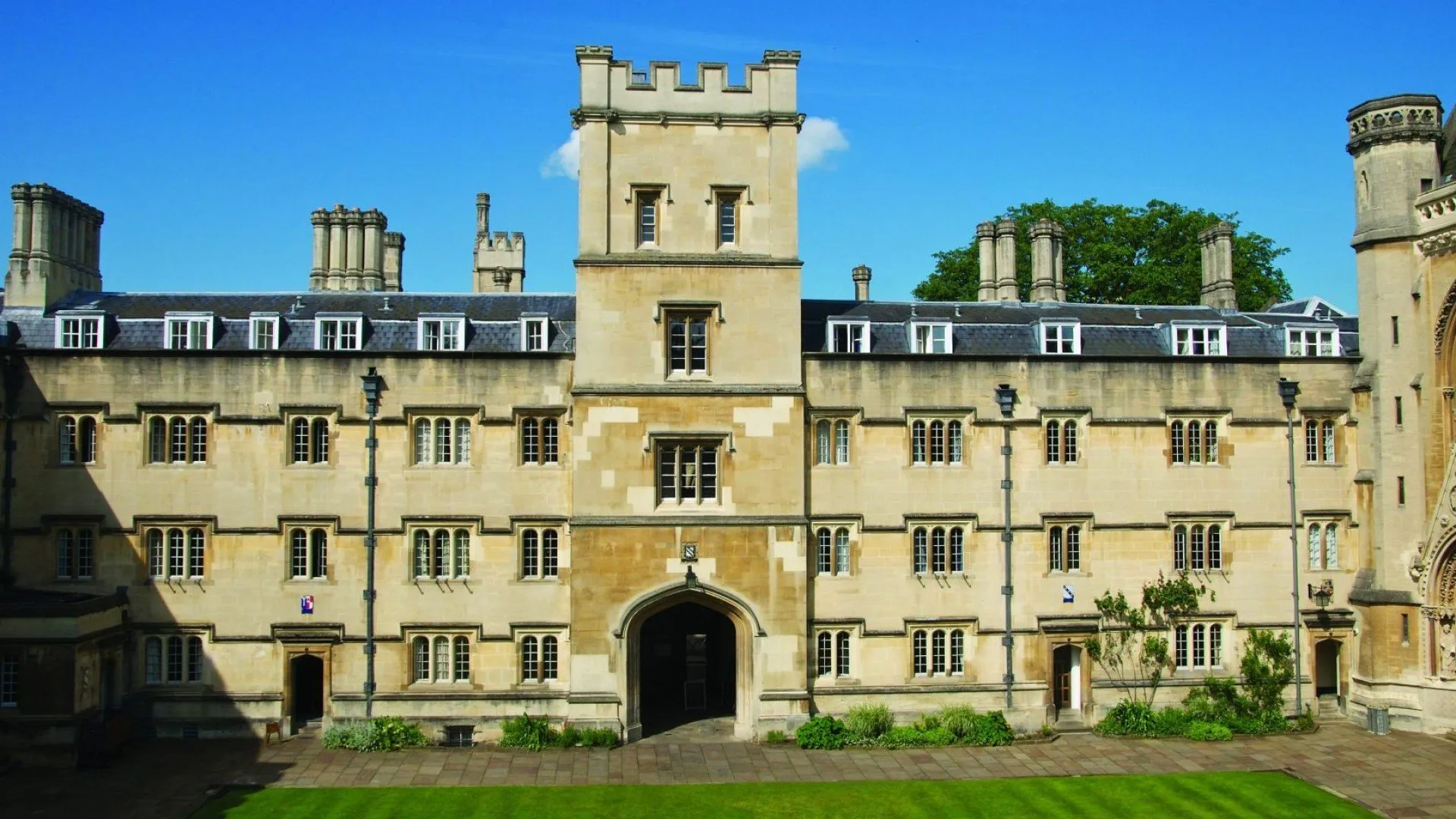 Kamienna fasada Exeter College w Oksfordzie z centralnie położoną wieżą z krenelażem, łukową bramą wejściową, rzędami okien dzielonych słupkami, kominami wzdłuż linii dachu oraz zielonym trawnikiem na pierwszym planie pod błękitnym niebem