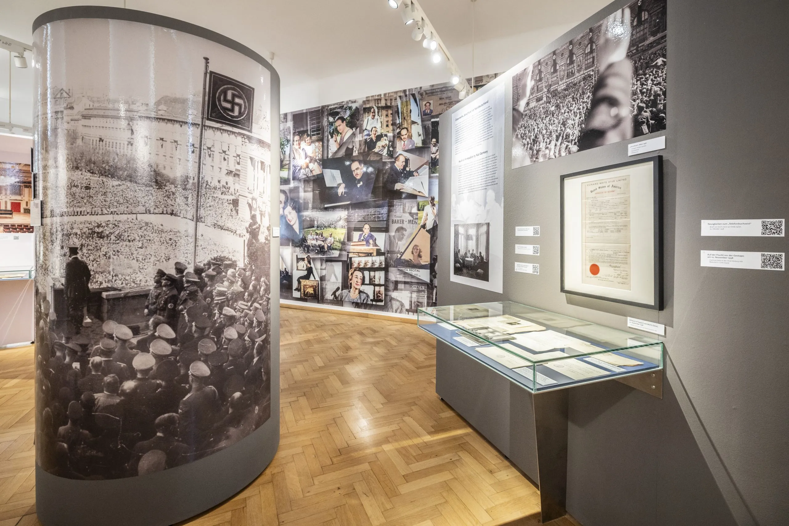 Wnętrze Centrum Exilarte z cylindrową gablotą prezentującą historyczne zdjęcie, ścianą czarno-białych obrazów oraz szklaną gablotą z dokumentami.