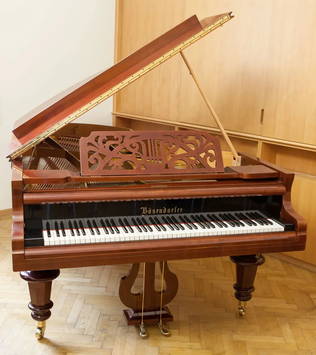Fortepian Bösendorfer z ozdobnym drewnianym pulpitem, otwartą klapą i polerowanym drewnianym wykończeniem, wyeksponowany wewnątrz na podłodze z parkietu.