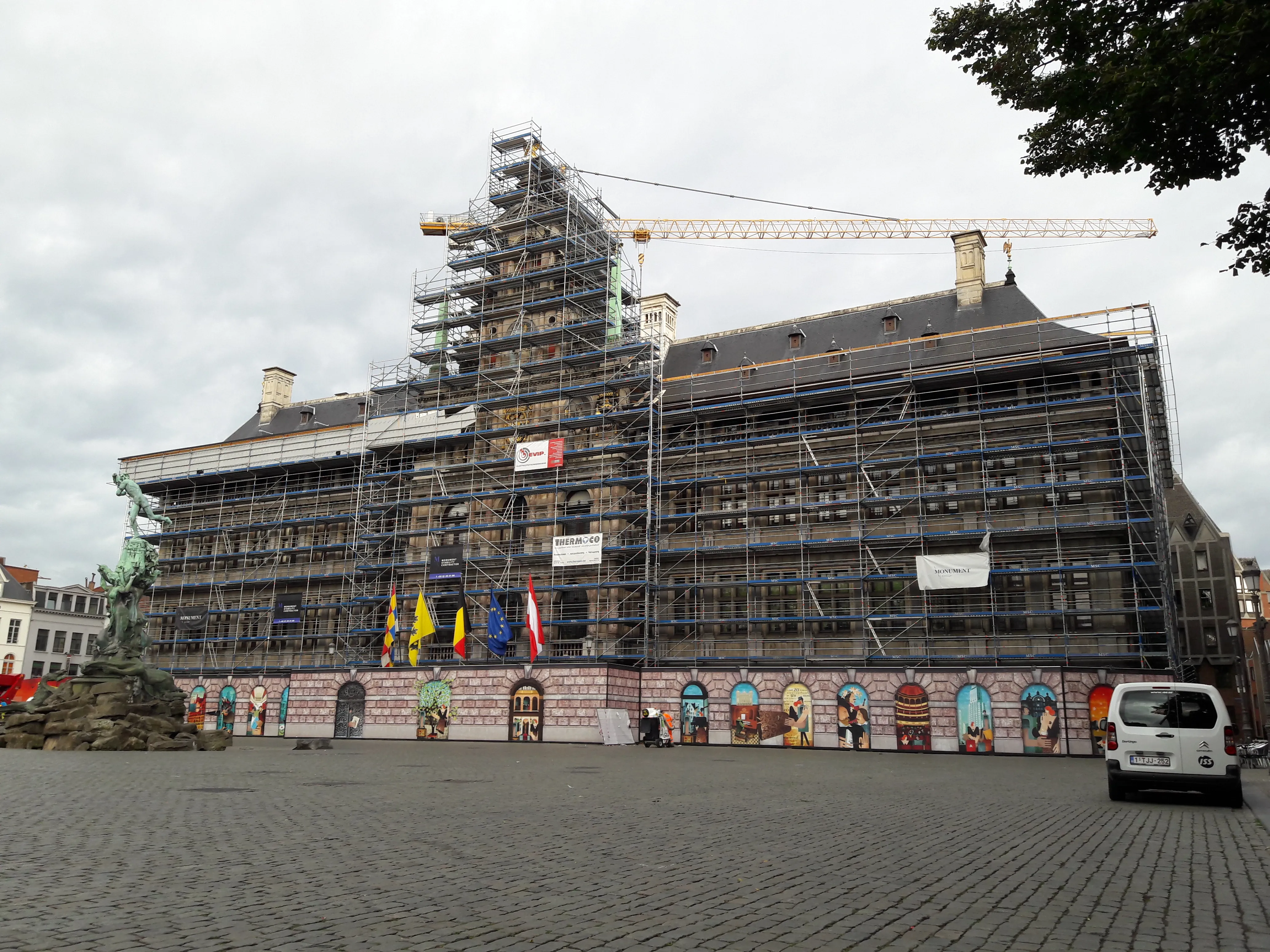 La fachada del Ayuntamiento de Amberes cubierta con andamios y una grúa de construcción durante su renovación, con banderas en la entrada y la fuente de Brabo visible a la izquierda