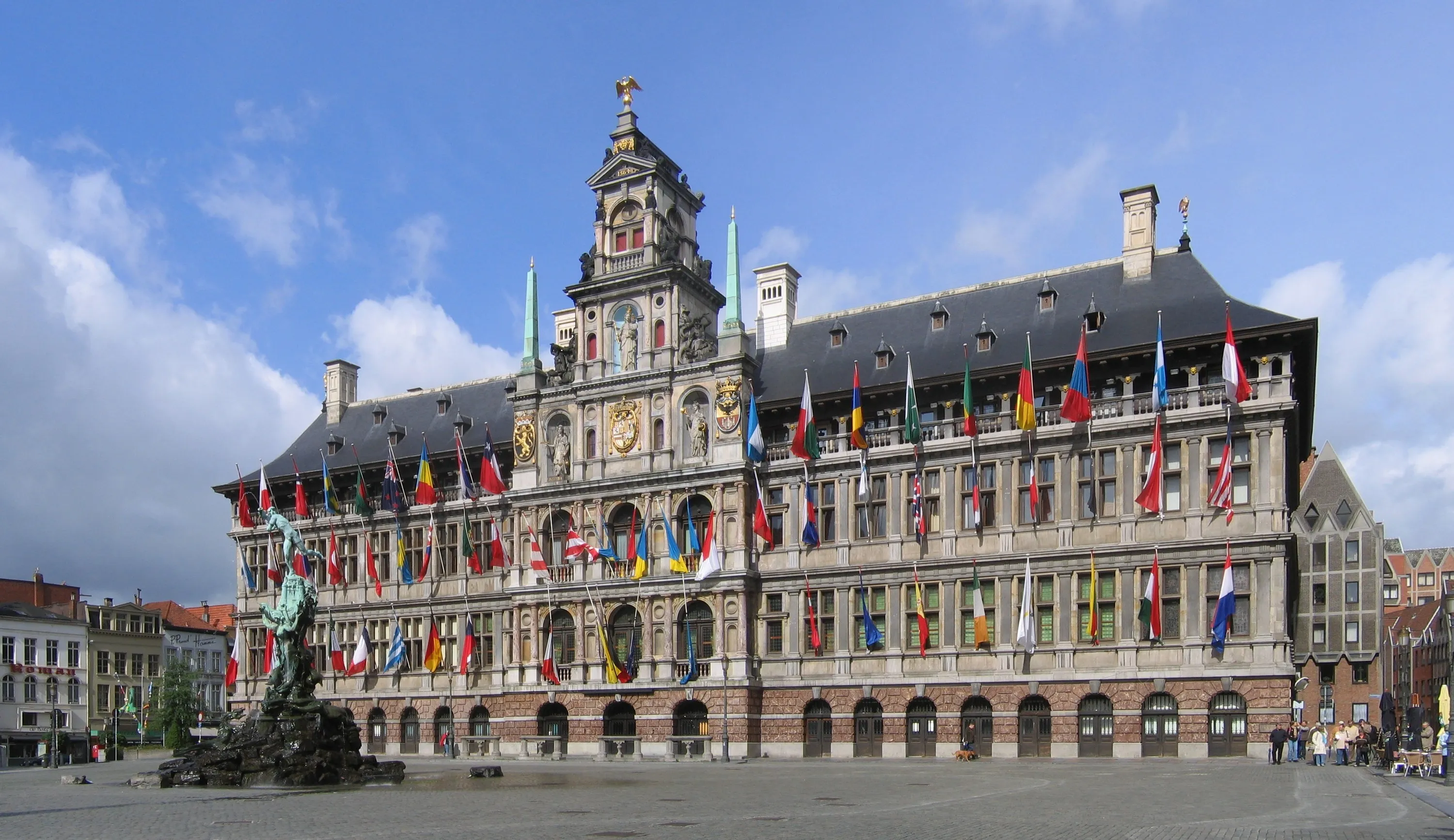 Vista exterior amplia del Ayuntamiento de Amberes con numerosas banderas internacionales colgando de la ornamentada fachada y la estatua de la fuente de Brabo en primer plano bajo un cielo azul con nubes.