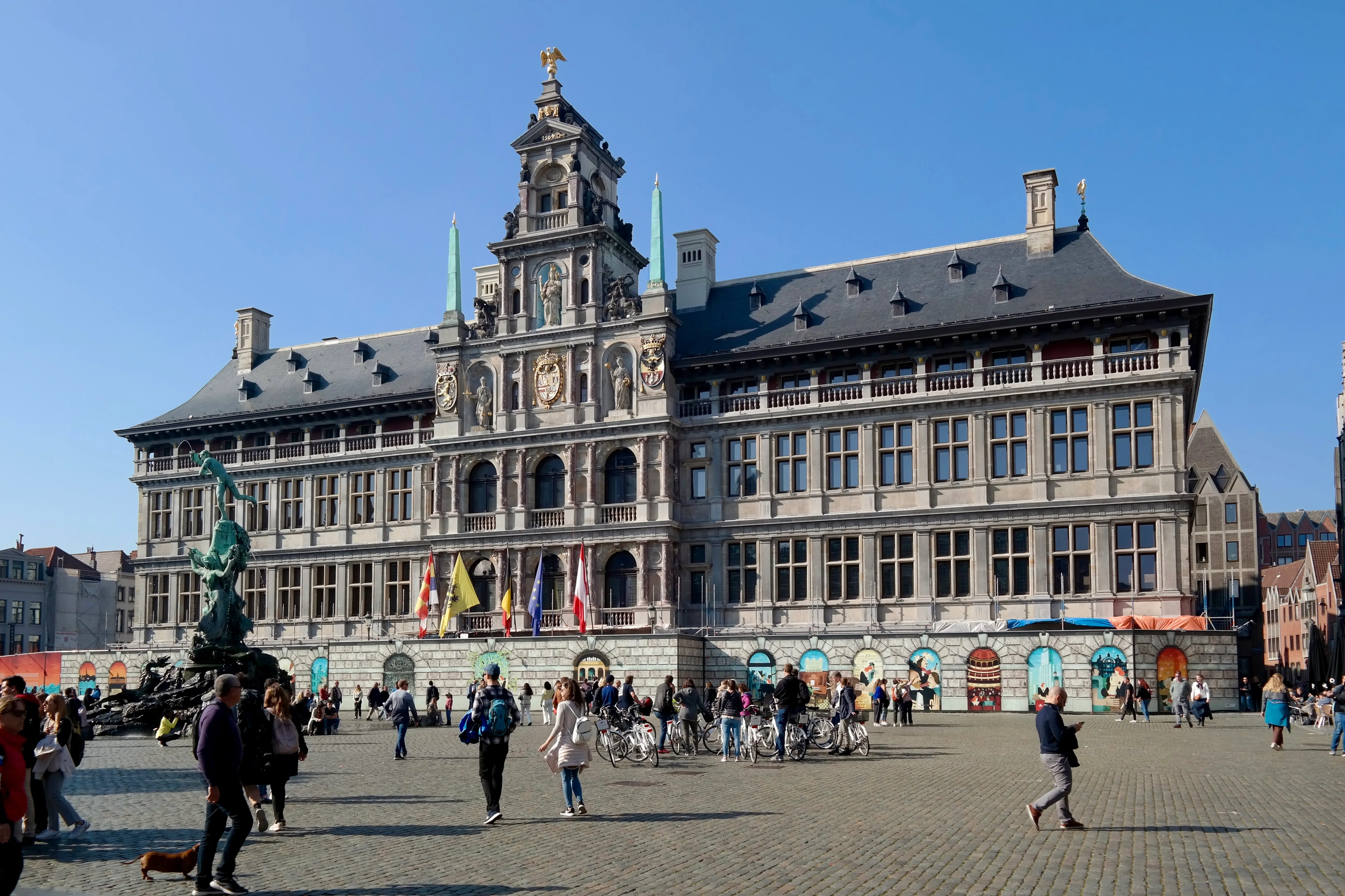 Vista exterior amplia de la fachada del Ayuntamiento de Amberes con su ornamentada arquitectura renacentista, torre del reloj y banderas, con gente caminando por la plaza empedrada frente al edificio
