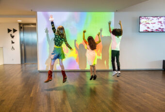Three children jump in front of a vibrant, multicolored projection on a large screen inside a museum with wooden flooring.