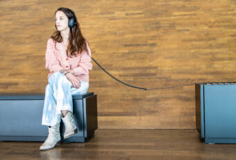 Woman sitting on a bench wearing headphones with a long cable extending to a display unit in a wooden-walled museum space.