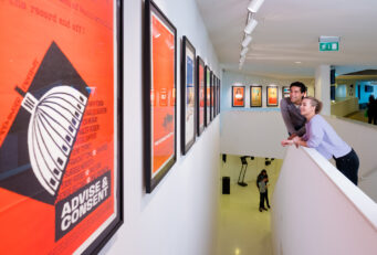 Modern museum hallway with orange-framed film posters, two people leaning on a railing overlooking a lower level with visitors