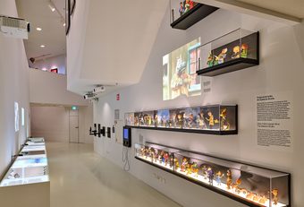 White-walled exhibition room with multiple glass display cases holding miniature film sets and a large screen showing a movie scene